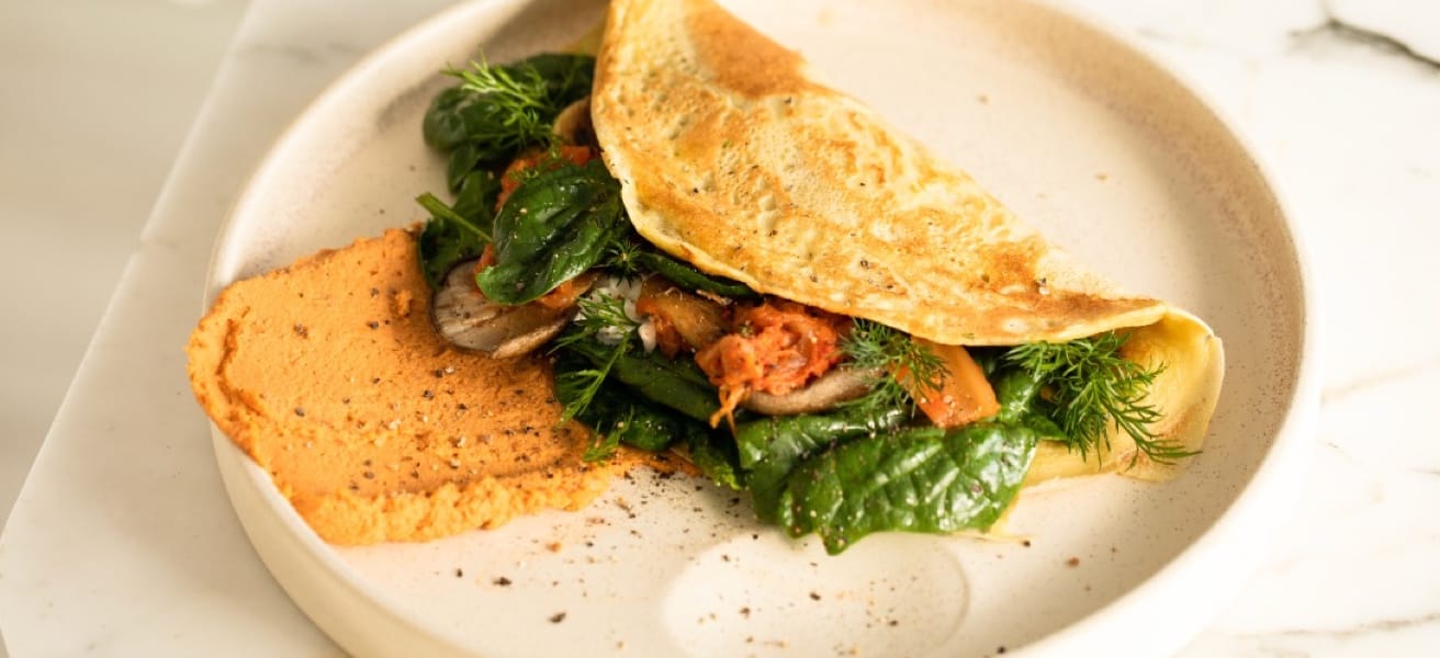 An image of a folded omelette on a circular plate stuffed with greens, herbs, and carrots on a sauce.